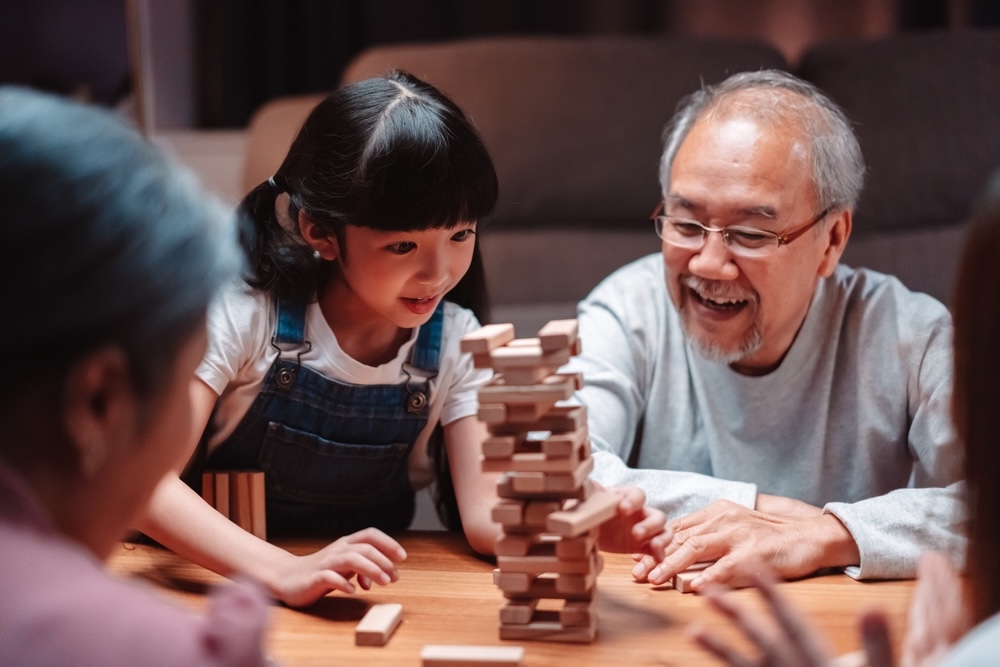 Grandparents and young girl having fun play stack tower wood block game at night in living room.