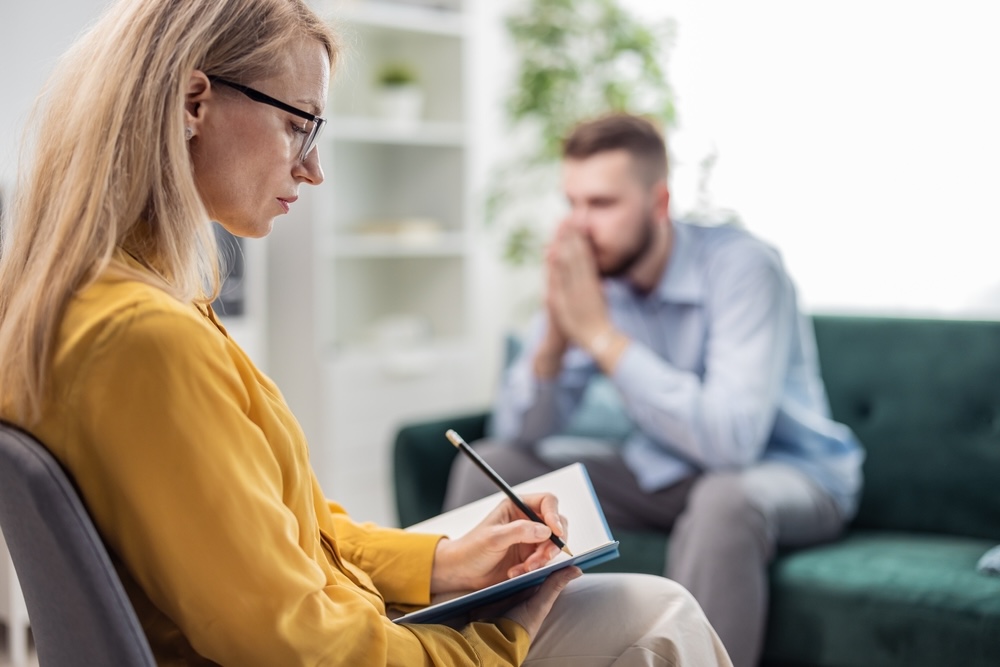 A psychologist making notes while listening to a male patient suffering from addiction.