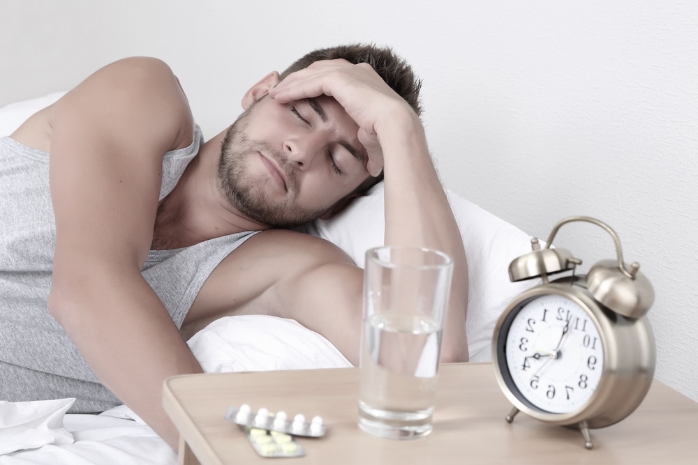 Man lying on the bed, experiencing a hangover, with medication, water and alarm clock on the bedside tab